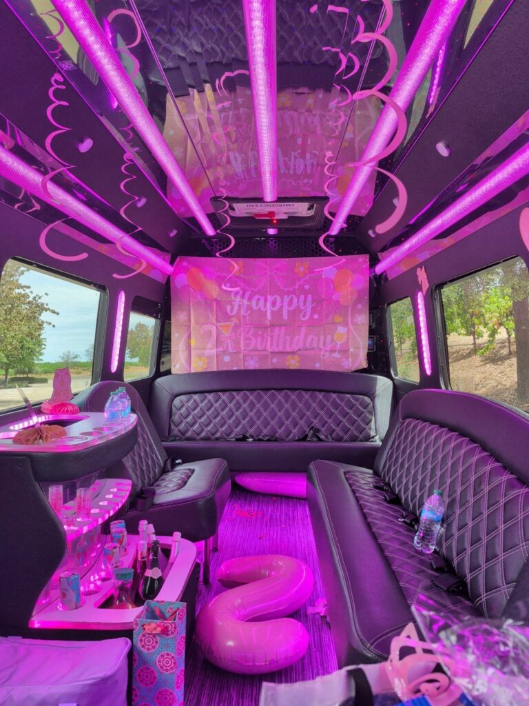 Party bus interior with pink neon lighting, black leather seats, and birthday decorations.