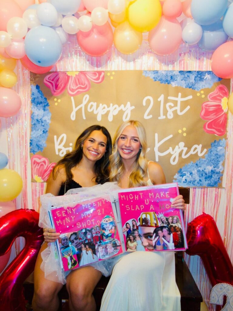 Two women holding a decorated 21st birthday shot book in front of balloons.