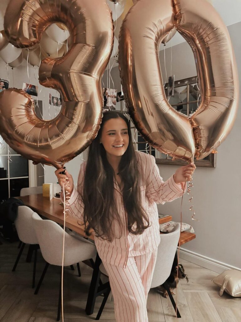 Woman in pink pajamas holding large rose gold balloons forming the number 30.