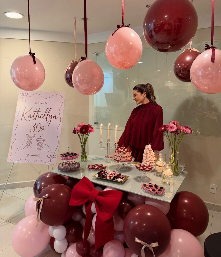 Woman standing behind a 30th birthday dessert table with burgundy and pink balloons.