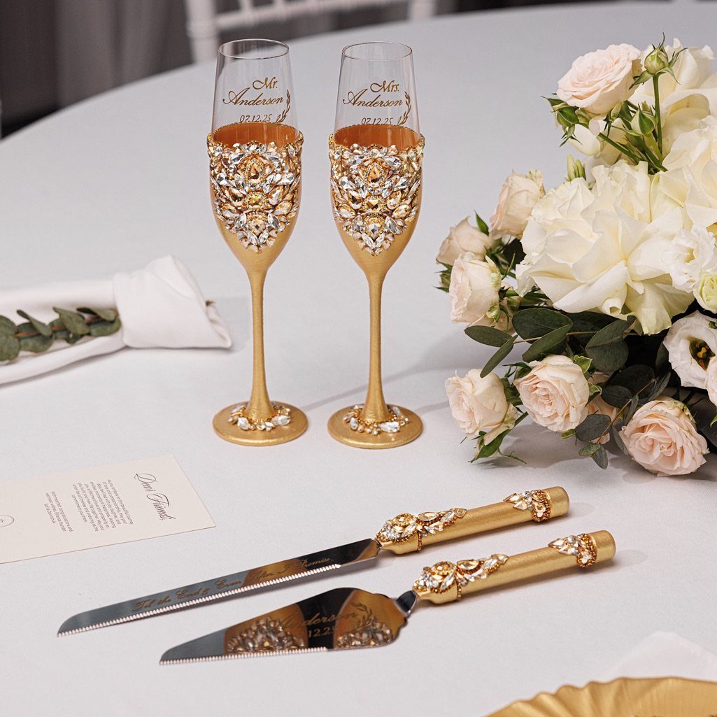 Two ornate gold and crystal champagne flutes beside a white rose bouquet.
