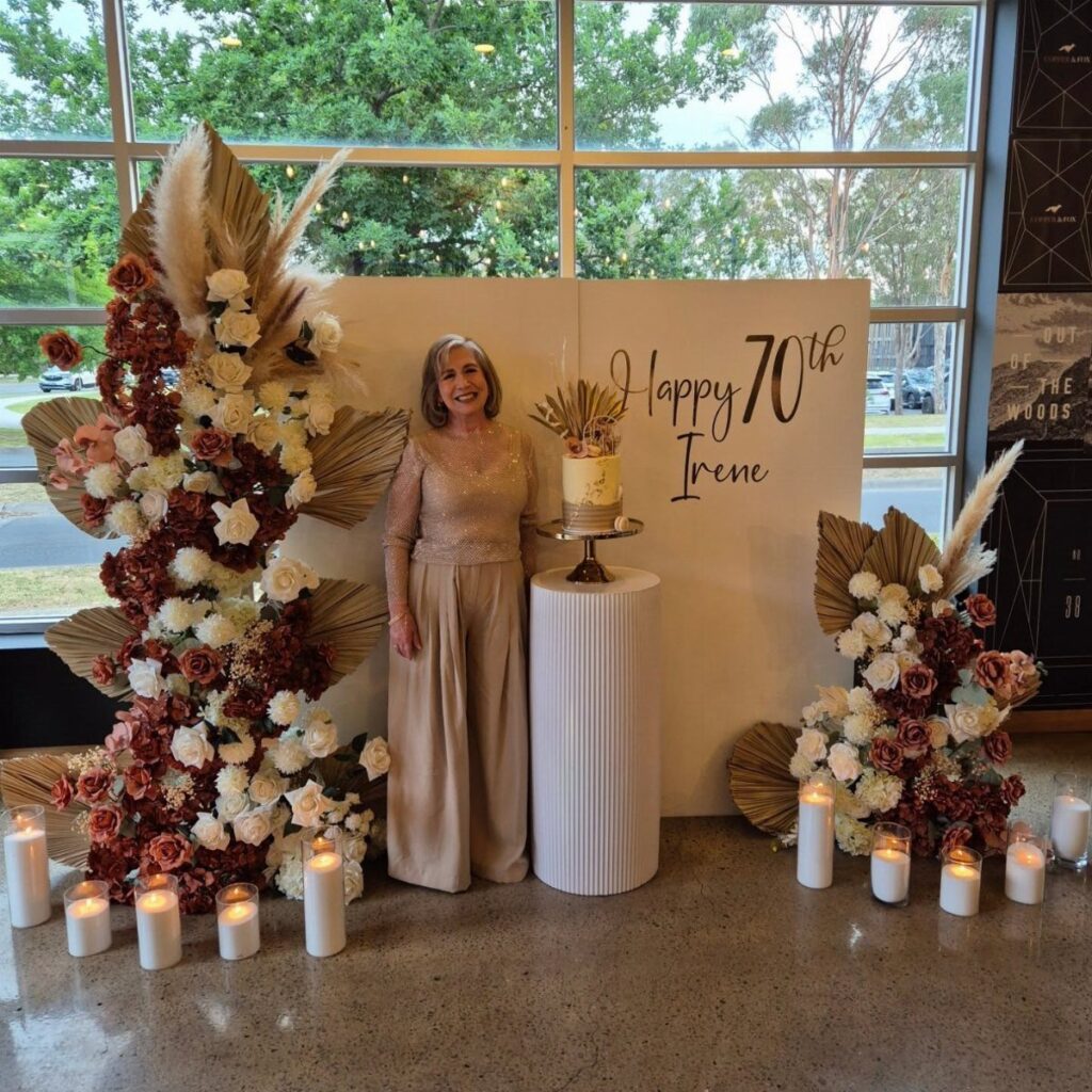 Woman celebrating her 70th birthday standing by a floral backdrop and cake.