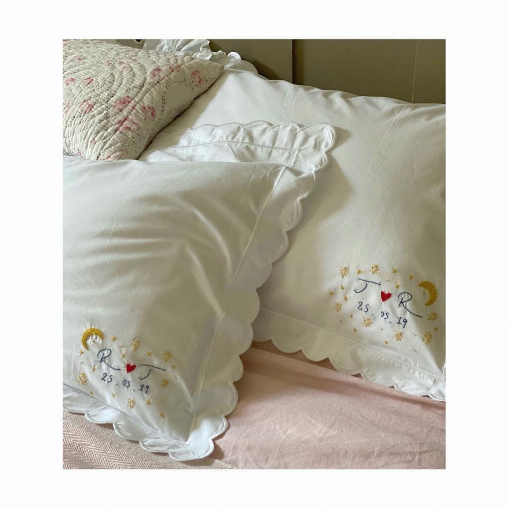 Embroidered white cotton pillowcases with initials and a date on a bed