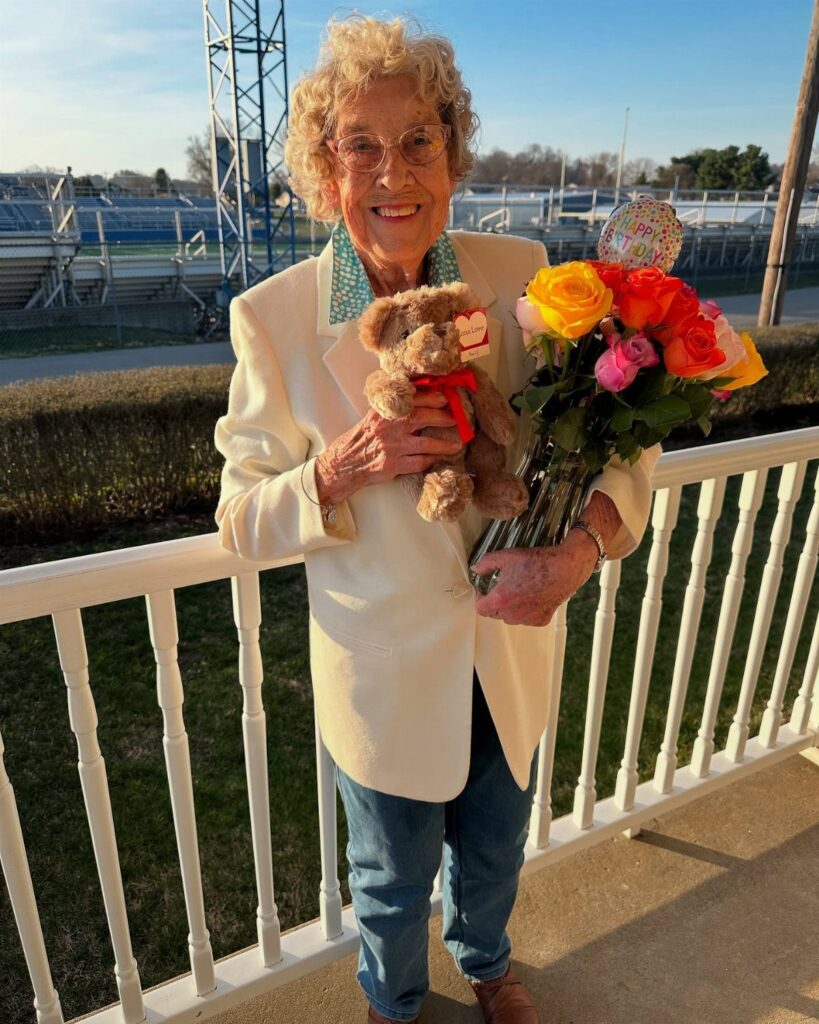 Elderly woman smiling and holding a teddy bear and colorful rose bouquet outdoors.