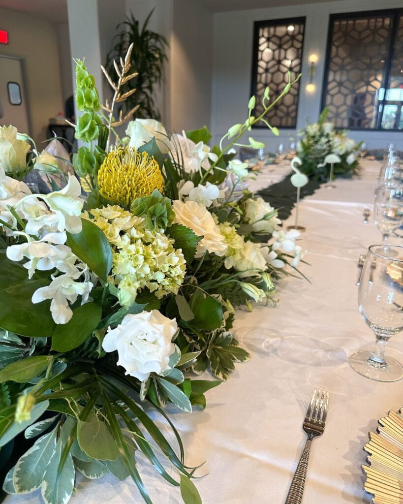White and yellow floral centerpiece with greenery on a white banquet table.