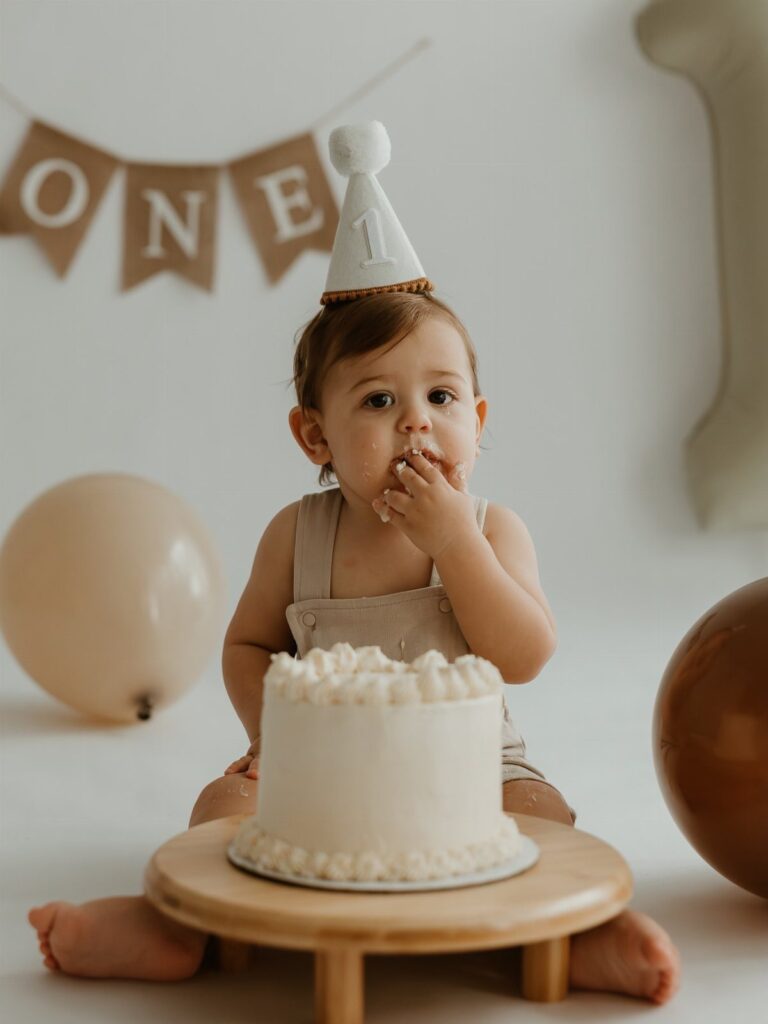 Baby in a party hat sitting with a white cake for a first birthday.