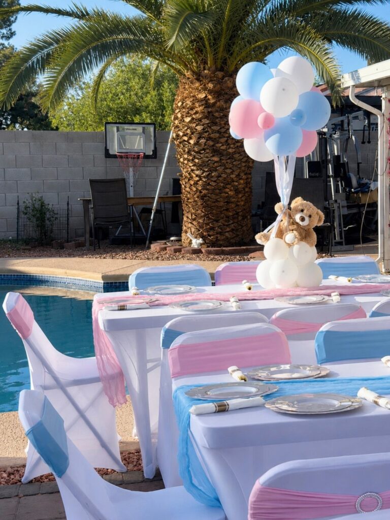 Outdoor party tables with pink and blue decorations, balloons, and a teddy bear.