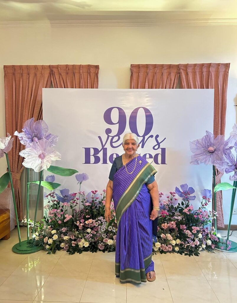 Elderly woman in purple saree posing before a 90th birthday celebration backdrop.