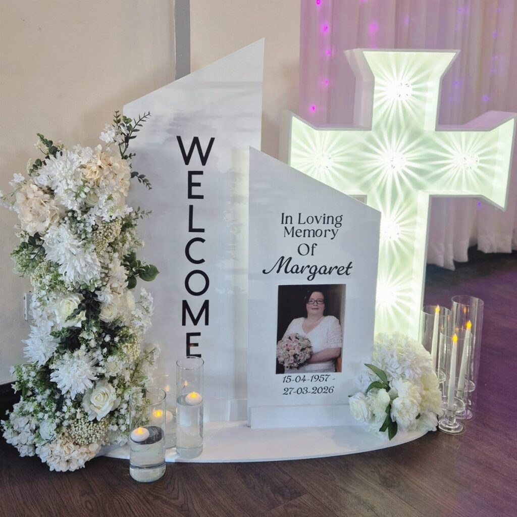 Memorial display with a photo board, illuminated cross, white flowers, and lit candles.