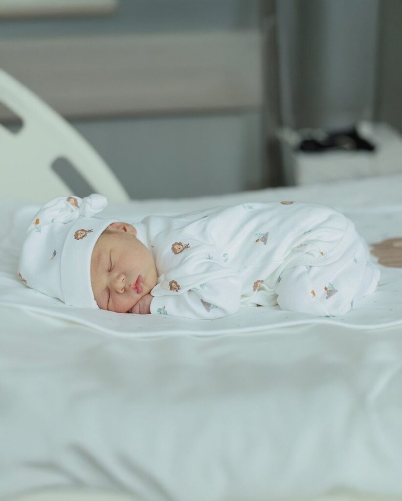 Sleeping newborn baby wearing a white patterned outfit and matching knotted hat.