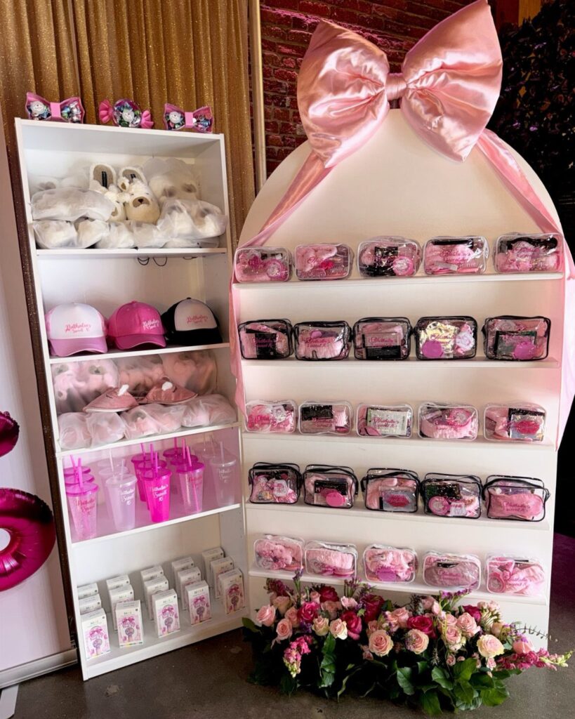 Pink and white party favors displayed on white shelves with a large pink bow.