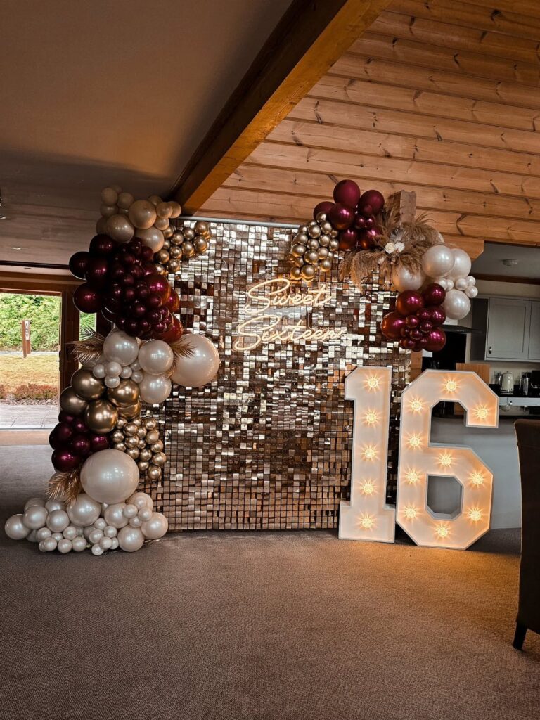 Gold sequin wall with burgundy and white balloons and glowing number 16